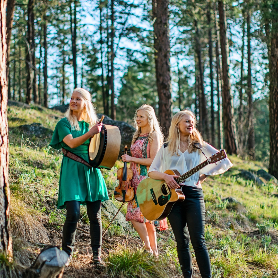 three sisters with blonde hair stand in the woods smiling and holding their instruments