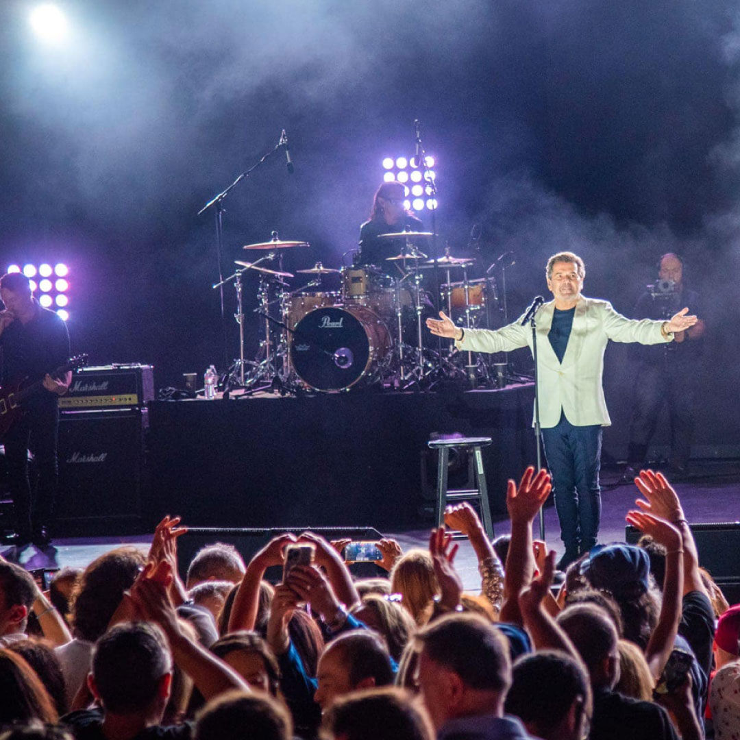 A man in a white jacket sings with his arms raised in front of a crowd